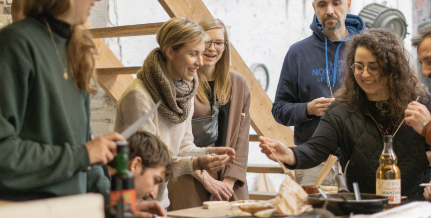 A group of five people gather around a table in the process of preparing tempeh. The people are smiling and handling indiscernible tools to prepare the tempeh