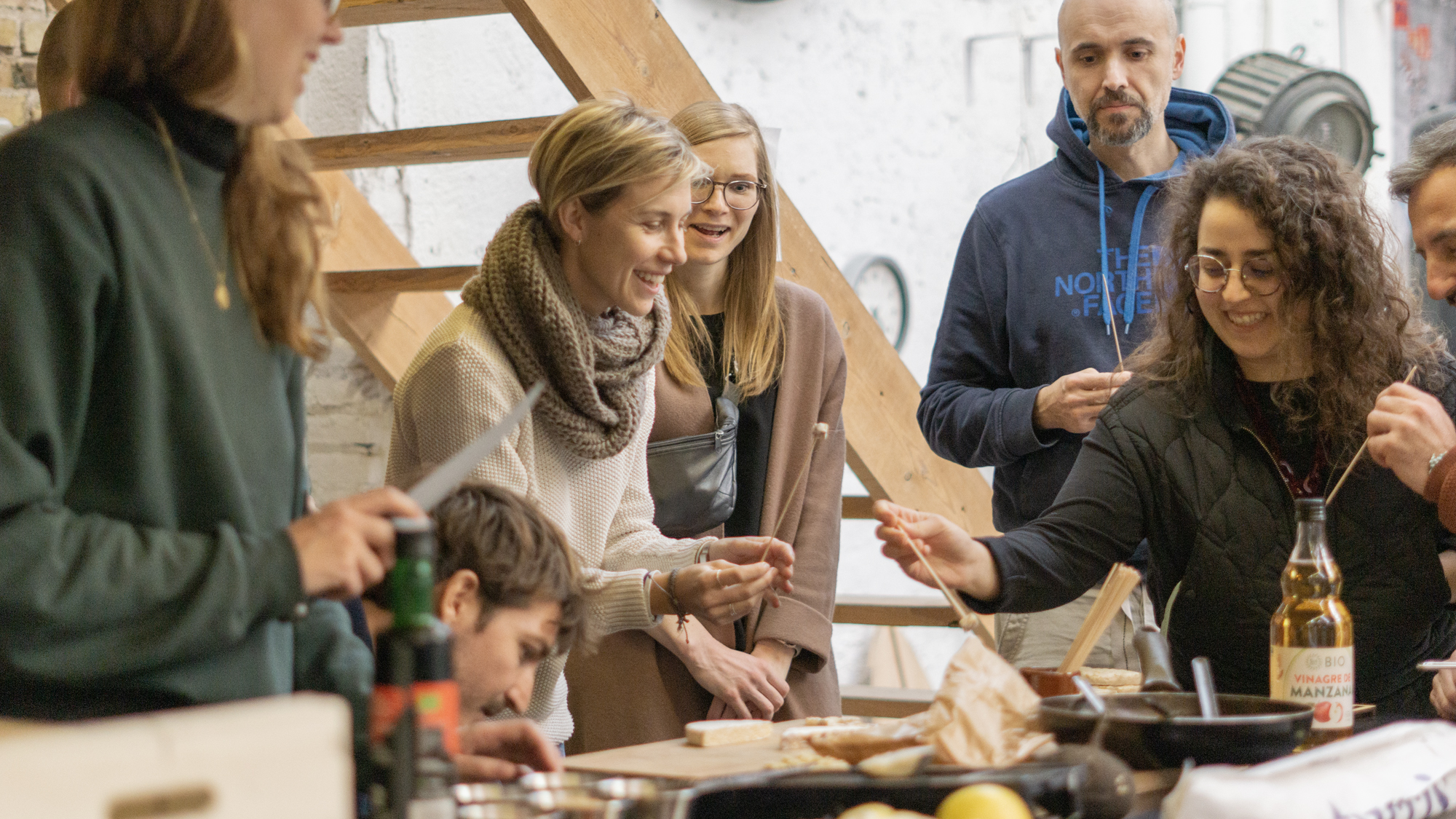 A group of five people gather around a table in the process of preparing tempeh. The people are smiling and handling indiscernible tools to prepare the tempeh