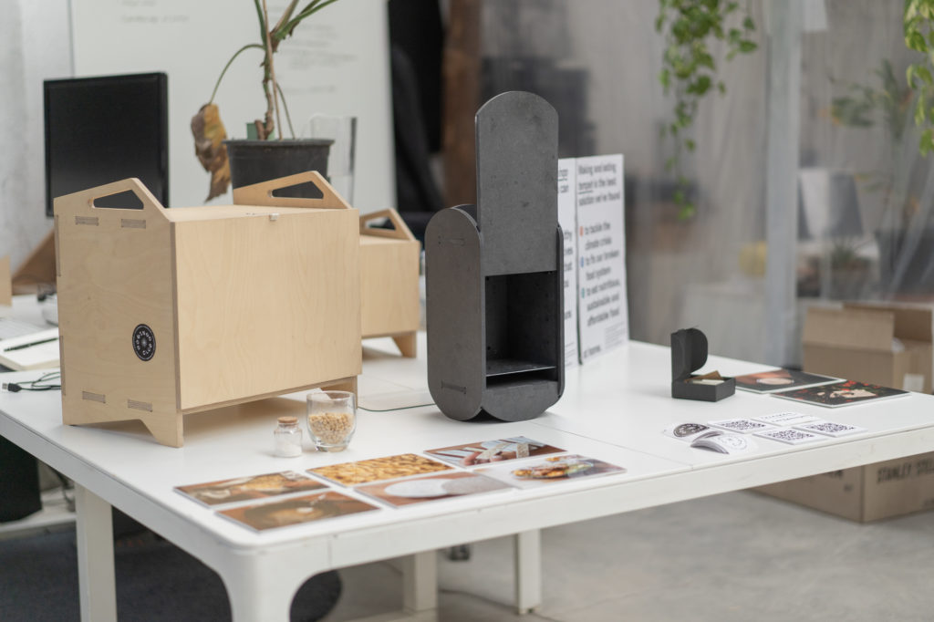 two incubators sit on a table. One is square and made of wood and the other is a relatively smaller rectangle made out of dark black material. The incubators sit behind images of tempeh