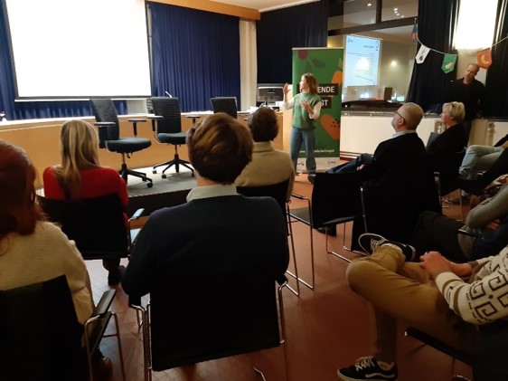 Workshop image in Ostend about a workshop related to food strategy directed to citizens - a room full of people sitting and visible from behind and at distance a woman standing and explaining the strategy. Behind her a roll-up poster with FoodSHIFT branding and elements is semi-visible