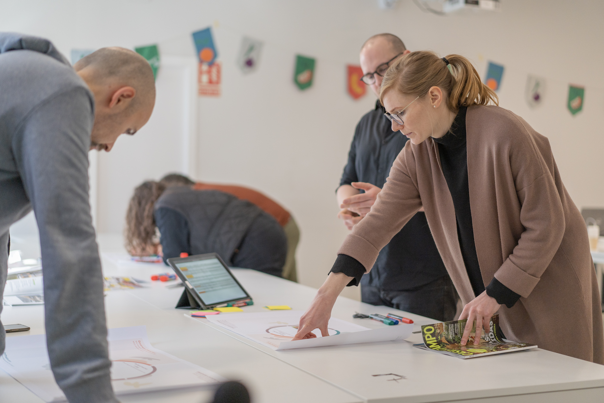 People in a room doing a workshop - a woman is pointing at a document on the table. Behind are more people and the FoodSHIFT2030 flags with their recognisable branding are hanging from the wall.